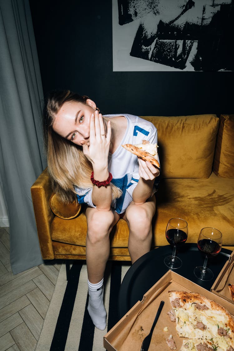 Young Woman Sitting On A Yellow Sofa While Eating Her Pizza