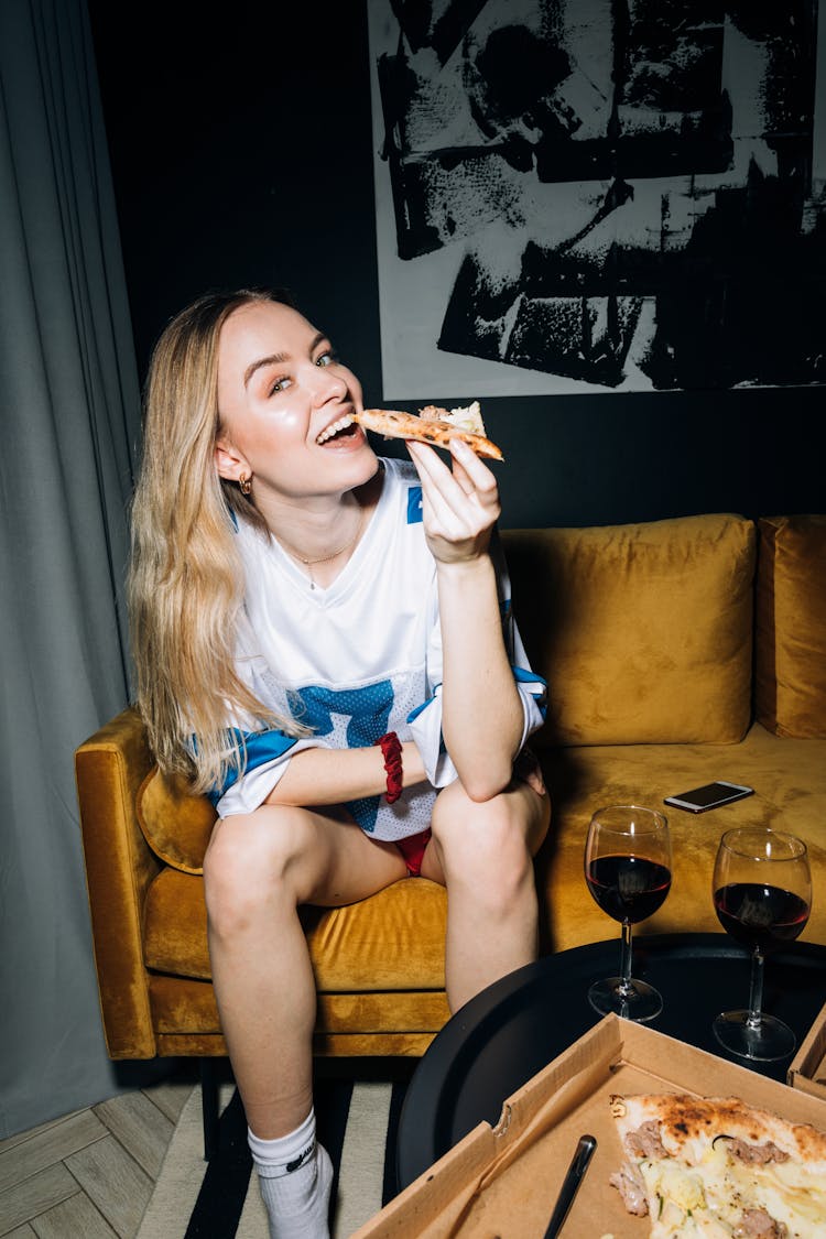 Young Woman Sitting On A Yellow Sofa While Eating Her Pizza