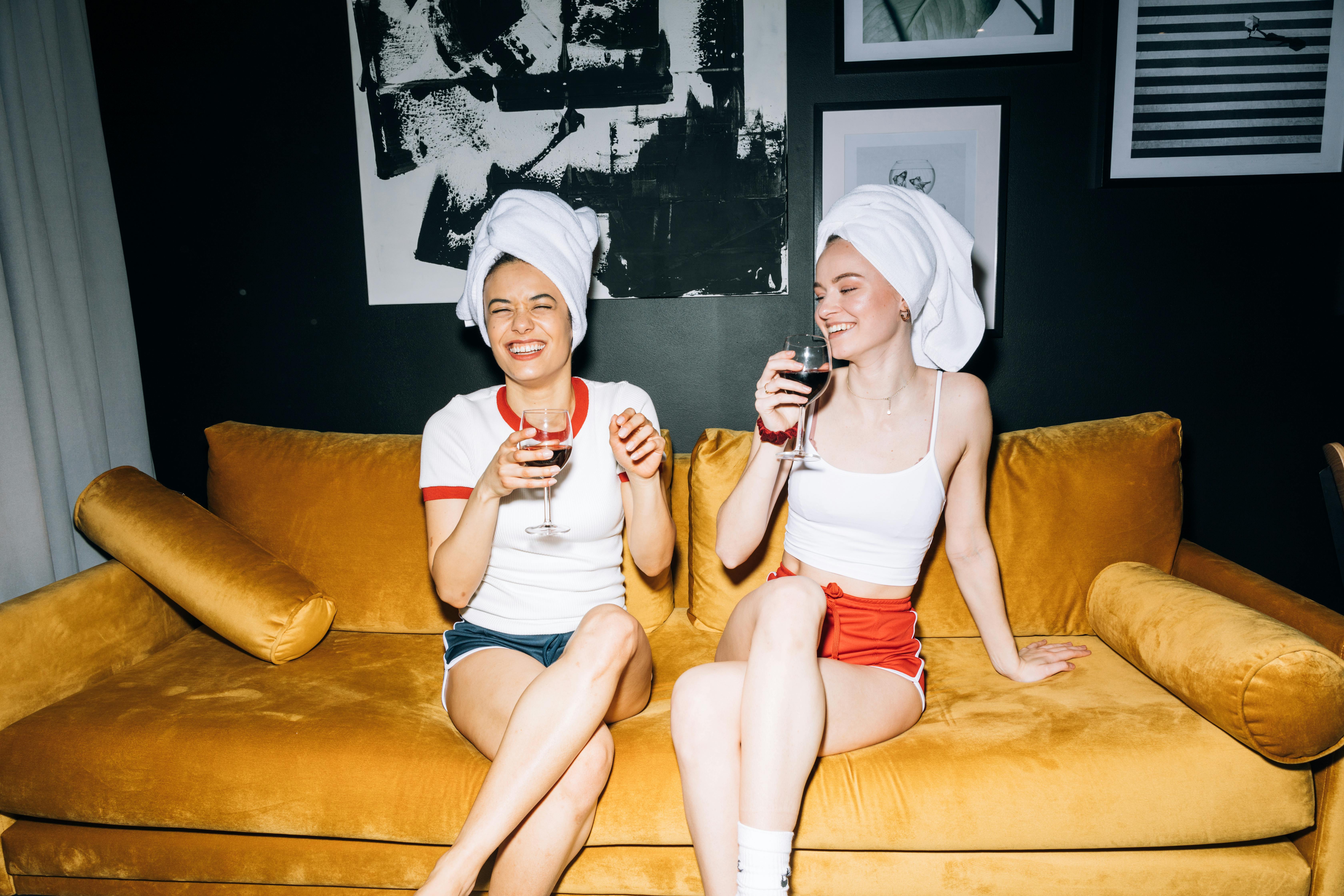 two young women sitting on a yellow sofa while holding a wine glass with red wine