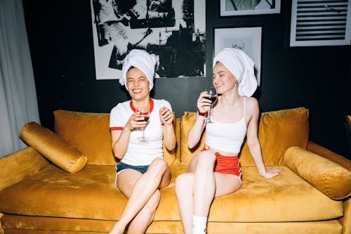 Free Two Young Women Sitting on a Yellow Sofa While Holding a Wine Glass With Red Wine Stock Photo