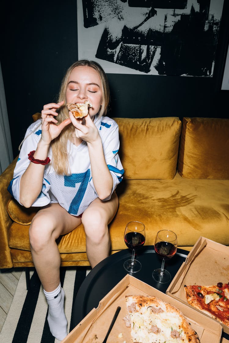 Young Woman Sitting On A Yellow Sofa While Eating Her Pizza