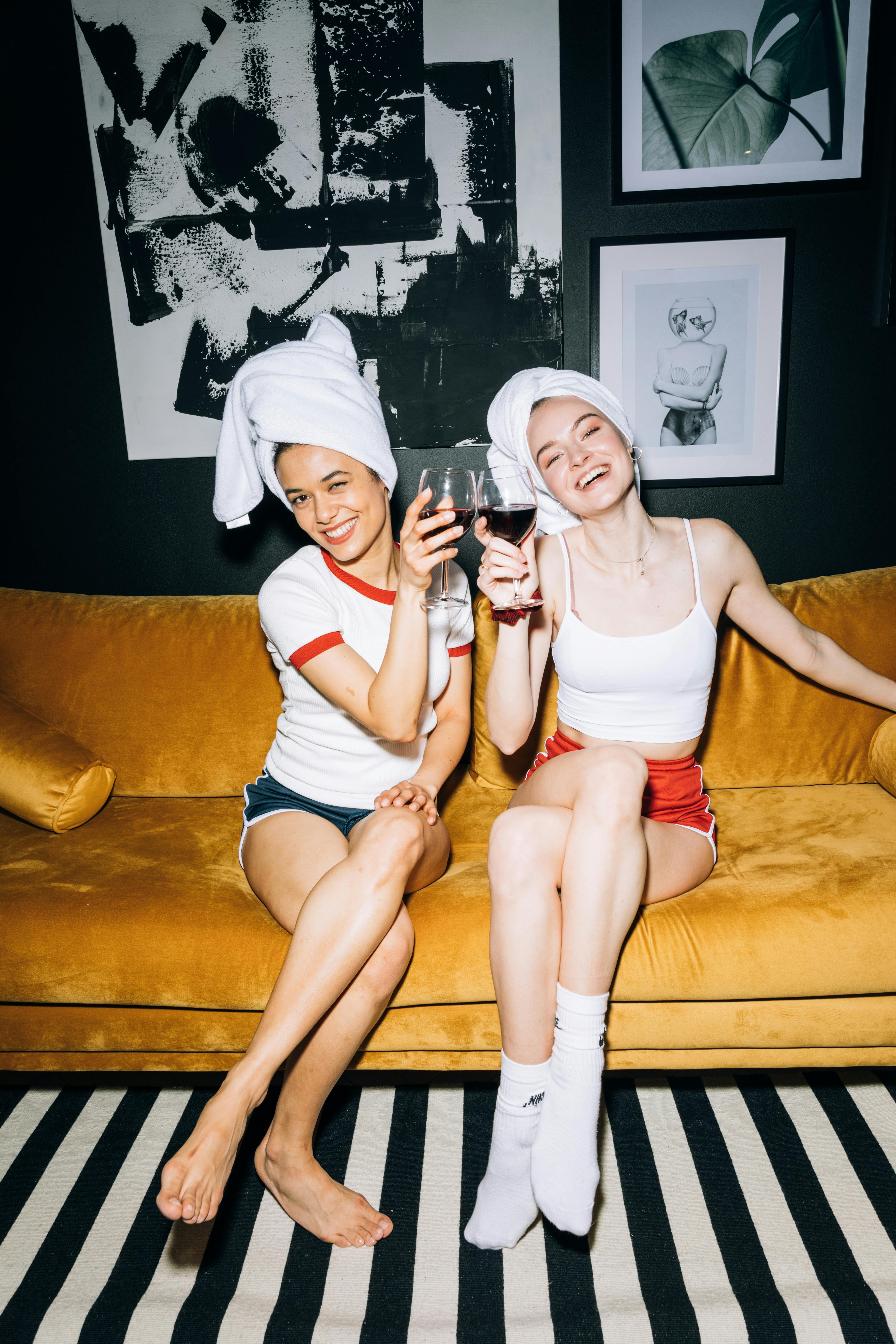 two young women sitting on a yellow sofa while holding a wine glass with red wine