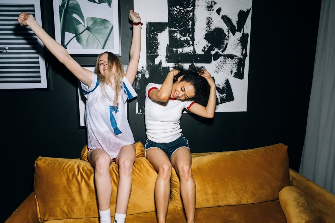 Two Young Women Having Fun While Sitting on a Yellow Sofa