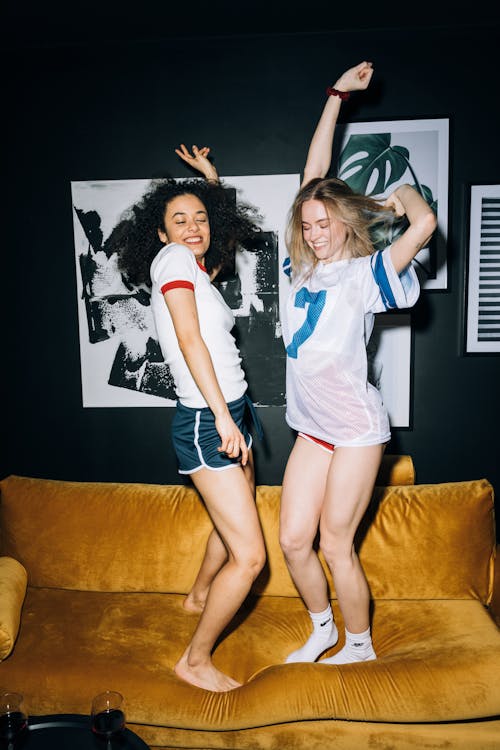 Two Young Women Having Fun Jumping on a Yellow Sofa