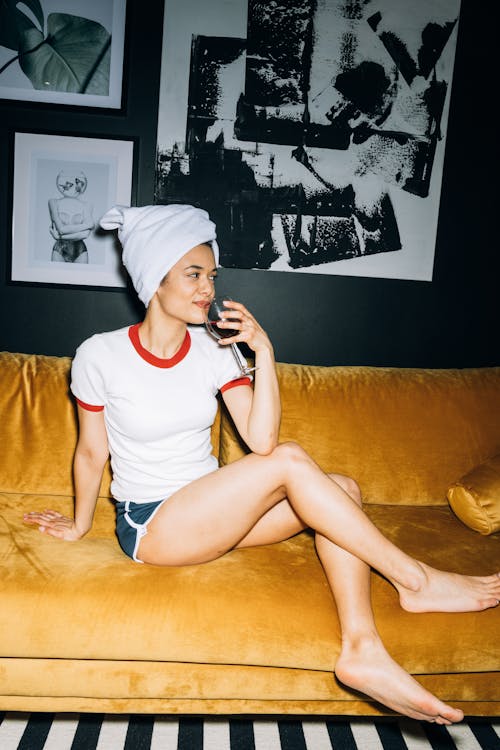 Free Young Woman Sitting on a Yellow Sofa While Holding a Wine Glass With Red Wine Stock Photo