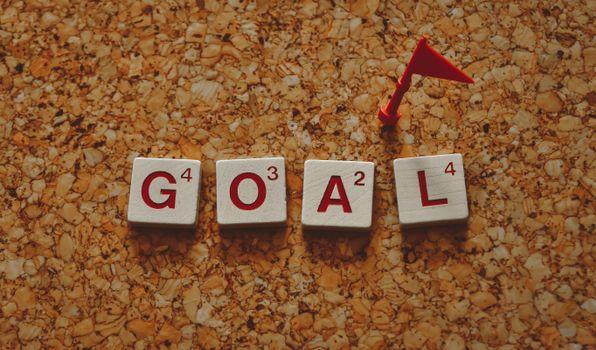 Scrabble tiles forming the word 'goal' with a small red flag on a cork board background.