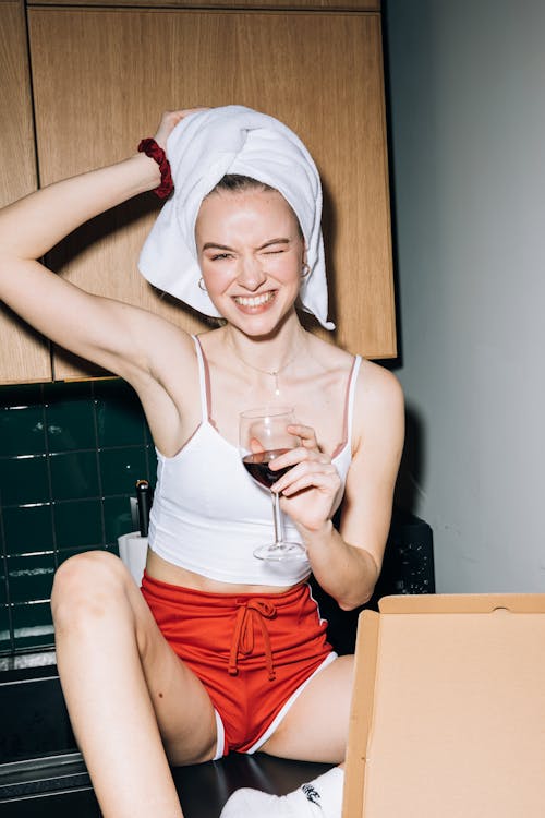 Free Young Woman in White Tank Top Smiling While Holding a Wine Glass With Red Wine Stock Photo