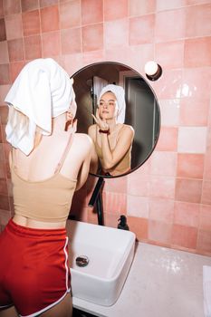 A young woman with a towel on her head enjoying her skincare routine in a stylish pink bathroom.