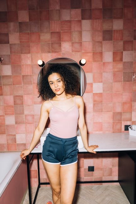 Young woman standing in a modern pink-tiled bathroom, exuding confidence and style.