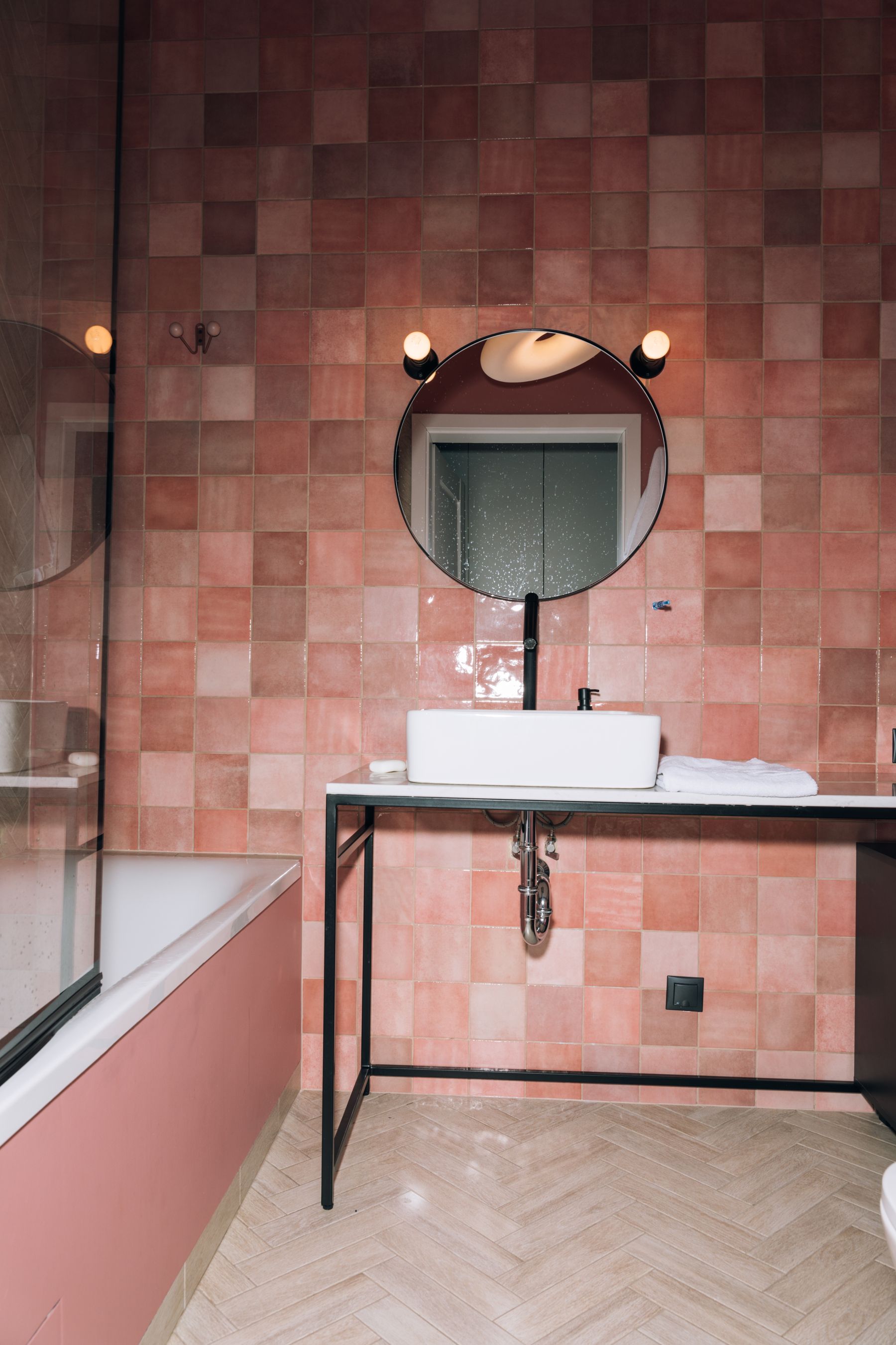 Clean pink bathroom with square-format pink tiles, matching bathtub, black-framed vanity and round mirror