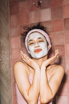 Young woman with a face mask in a pink bathroom, enjoying her skincare routine with a joyful expression.