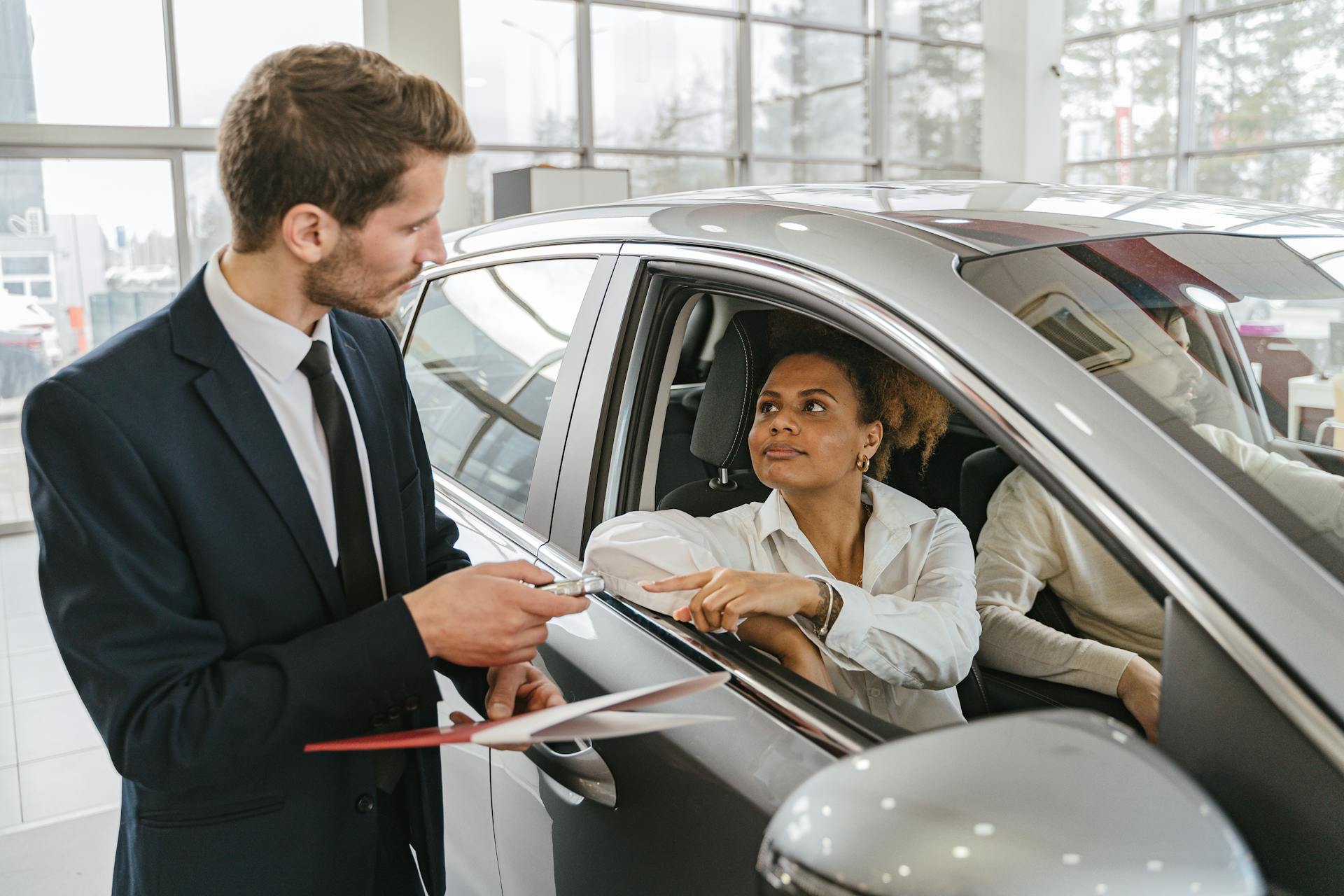 A car dealer hands keys to a woman sitting inside a vehicle in a car showroom.