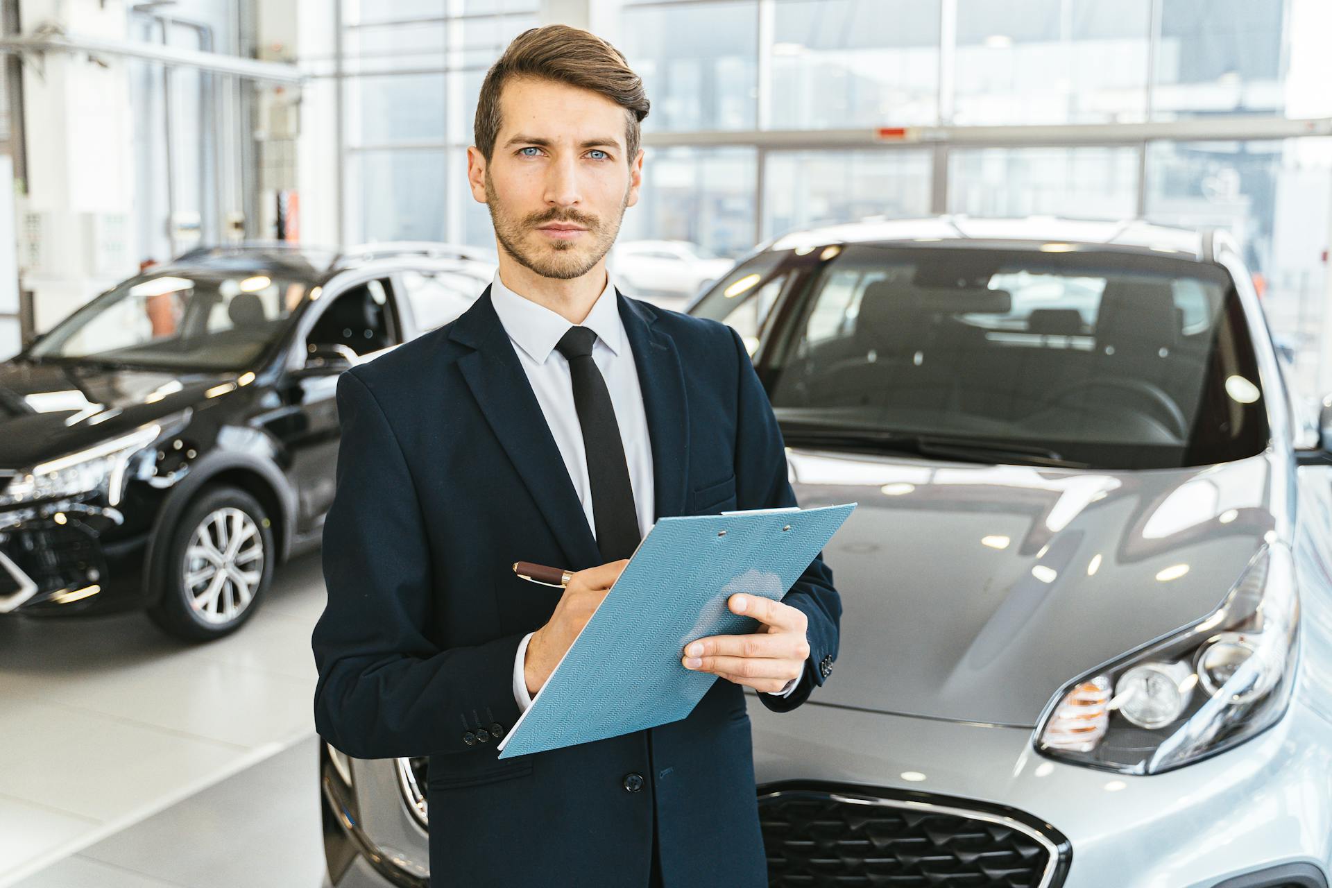 A confident car salesman in a showroom, holding a clipboard and pen.