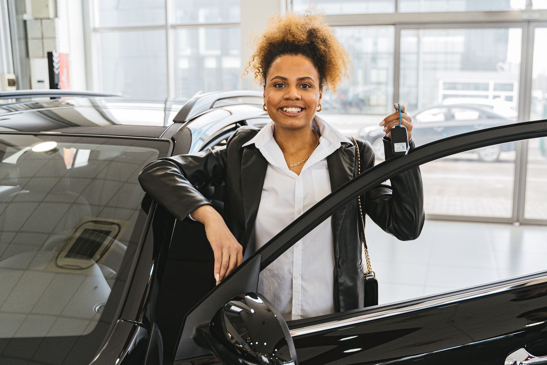 Smiling woman with car keys standing beside her new vehicle in a dealership showroom.