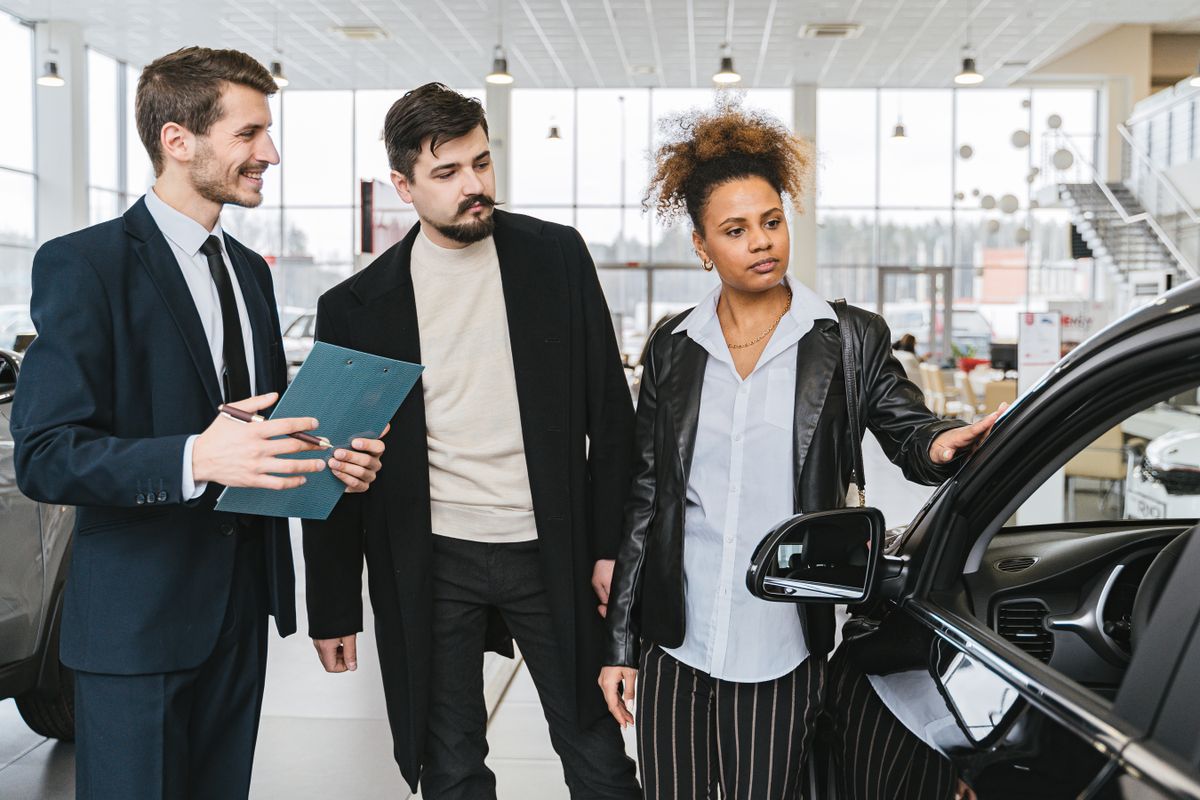A smiling group at a car dealership reviewing paperwork next to a vehicle