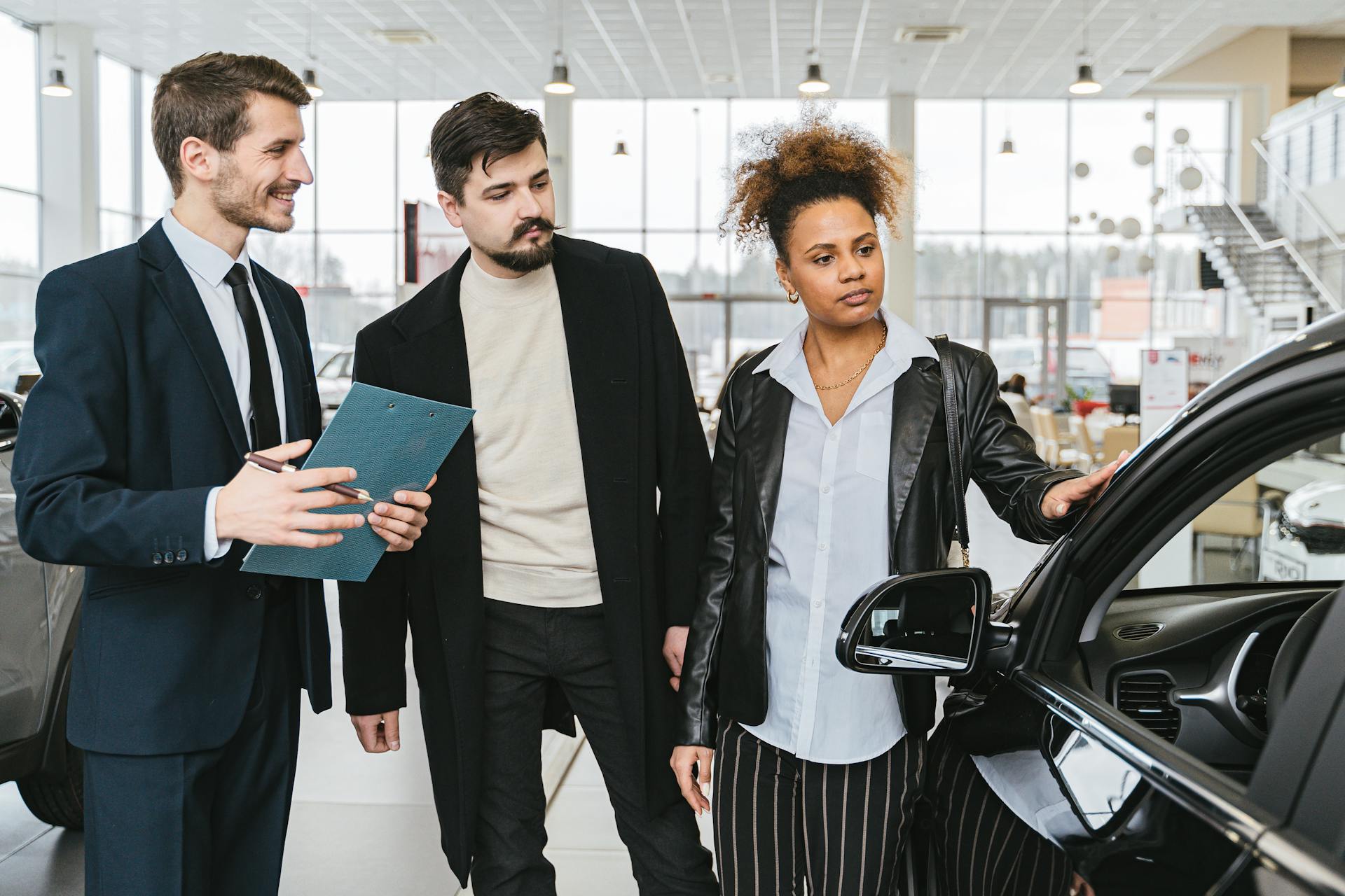 An interracial couple consults with a salesman at a car dealership, exploring vehicle options.