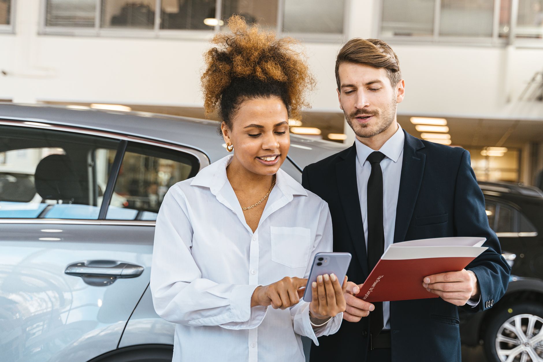 Two professionals discussing business while looking at a mobile phone in a car showroom.