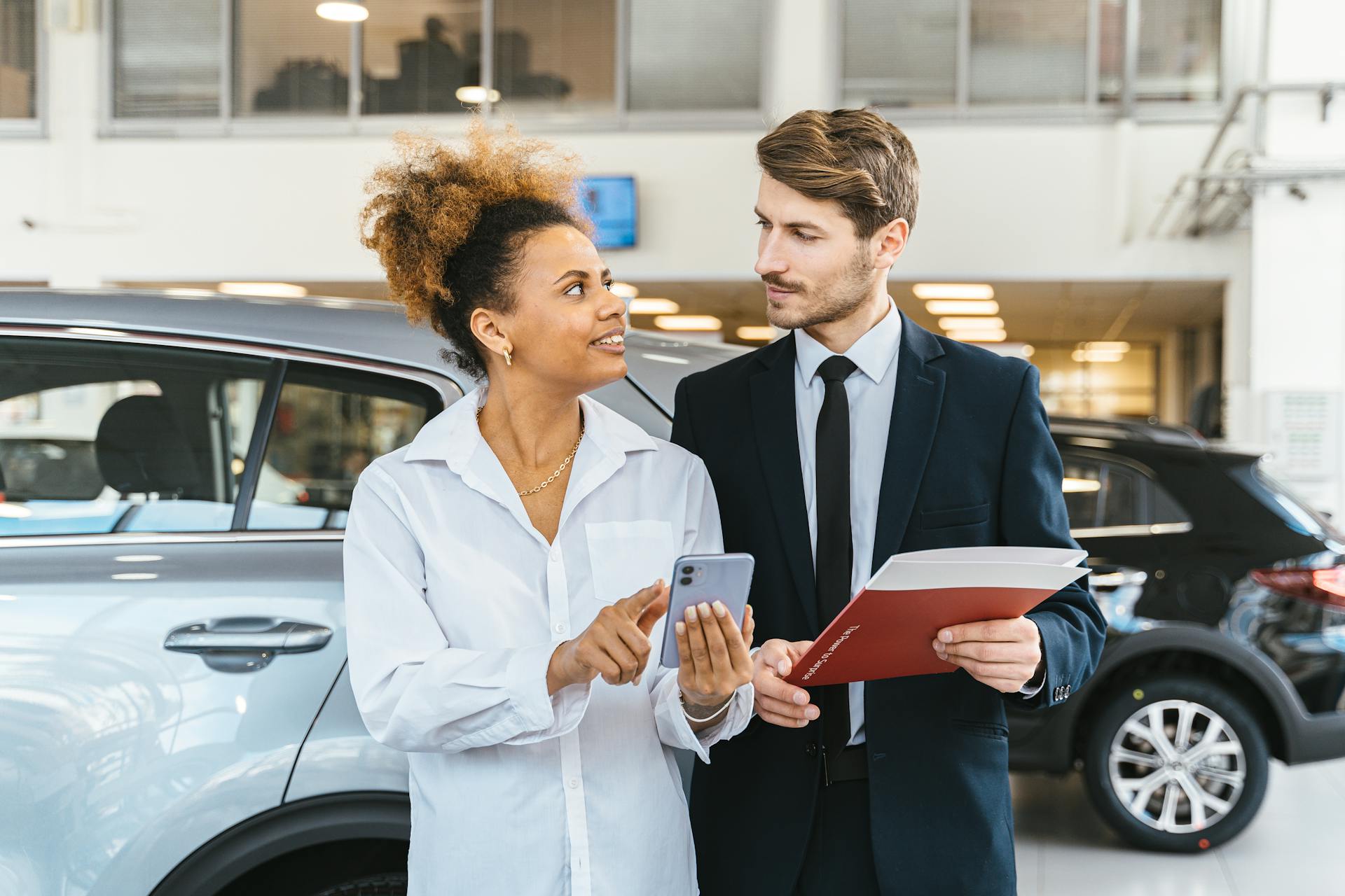 African American woman and Caucasian man discuss car purchase at dealership using smartphone.