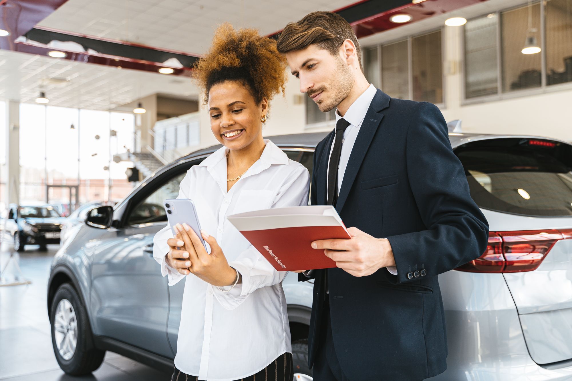 Shows a customer and salesperson at a car dealership with a phone screen visible, closely matching the…