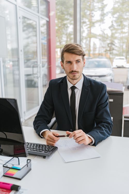 Free Professional businessman in a suit sitting at a desk in a modern office, holding a pen with documents. Stock Photo