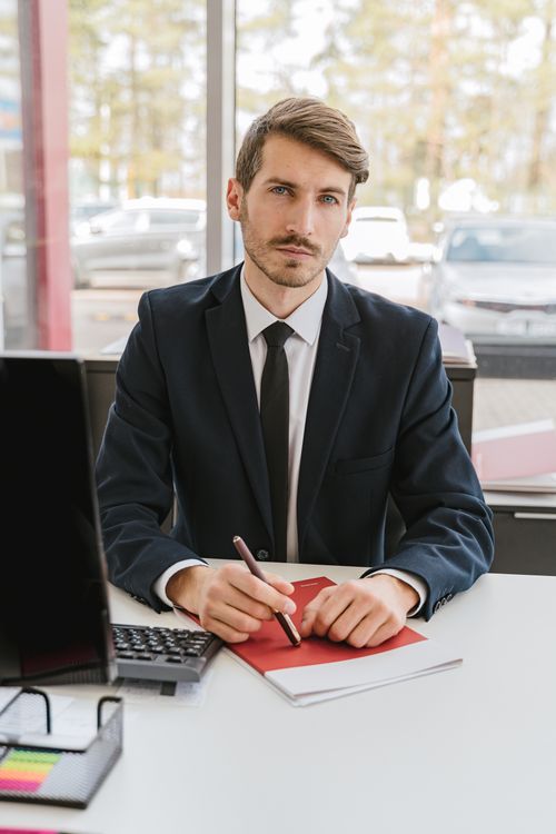 Free Professional businessman in suit sitting at a desk, pen in hand, serious expression, office environment. Stock Photo
