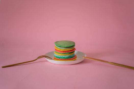 Tasty multicolored pancakes served on white plate for breakfast on pink background of studio