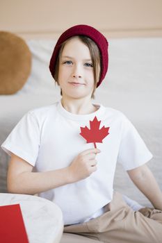 Child wearing a beanie holds a red maple leaf symbol, celebrating Canada Day indoors.