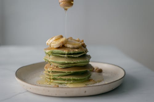 Free Unrecognizable person pouring honey on stack of appetizing green pancakes topped with bananas and walnuts and served on plate on table Stock Photo