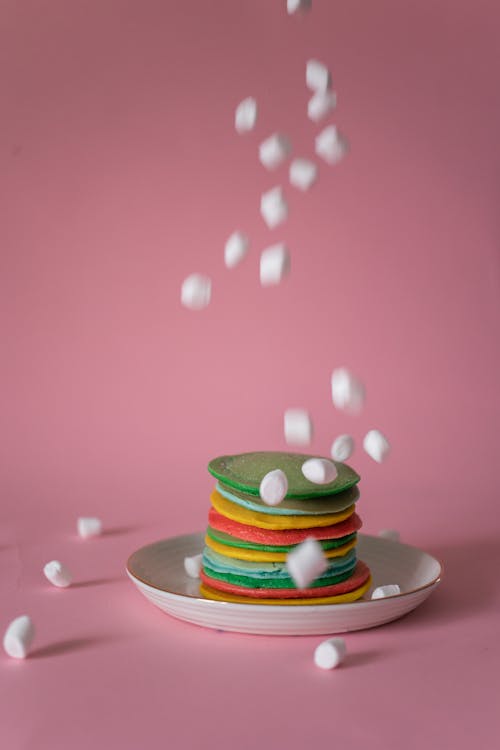 White marshmallows falling on plate with stack of colorful pancakes placed against pink background in studio