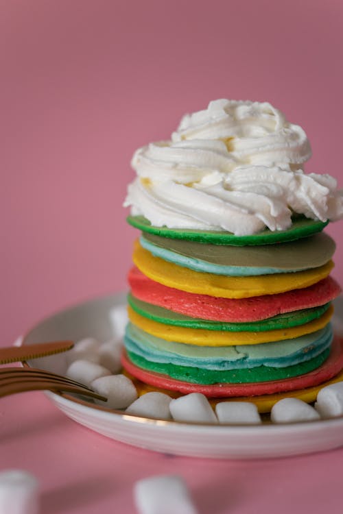 Ceramic plate with stack of multicolored delicious pancakes decorated with whipped cream and marshmallows in pink studio
