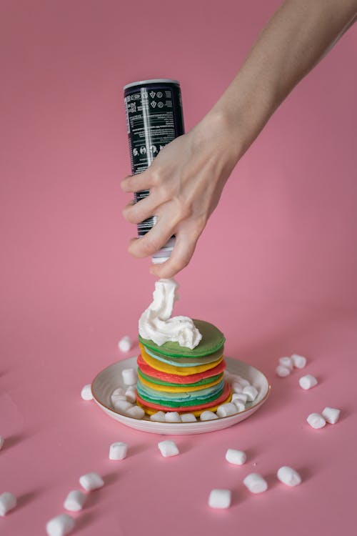 From above of crop unrecognizable female decorating stack of colorful freshly prepared pancakes with whipped cream spray against pink background