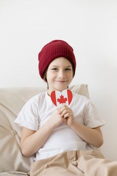 A cheerful child in a red beanie holds a heart-shaped Canadian flag indoors.