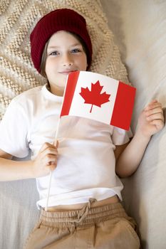 A smiling child lying down while holding a Canadian flag indoors.