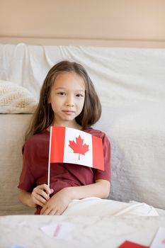 Young girl smiling while holding a Canadian flag, celebrating Canada Day indoors.