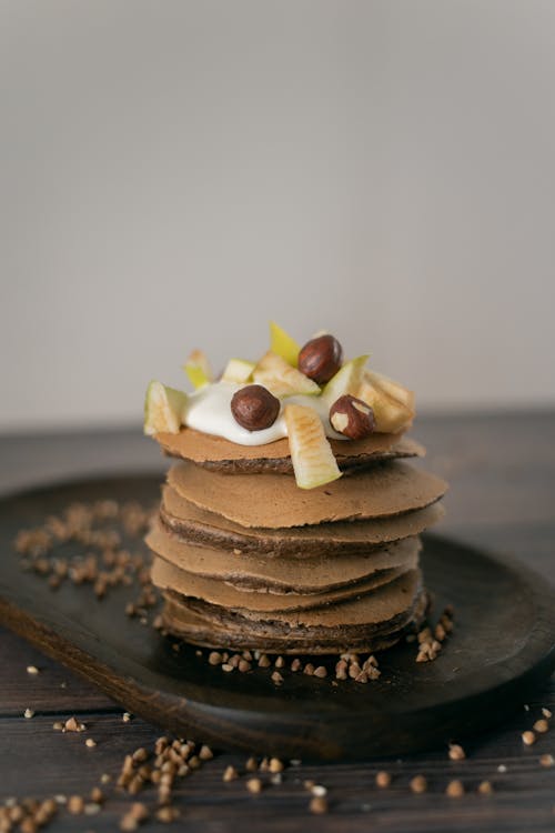 High angle of delicious homemade chocolate pancakes decorated with fruits and hazelnuts and served on plate with scattered buckwheat grains