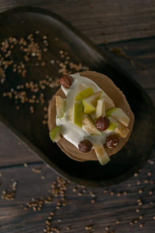 Top view of delicious pancakes topped with cream and decorated with hazelnuts and sliced apples on plate served on wooden table