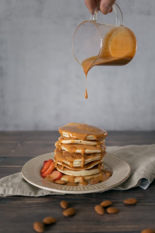 Free Crop anonymous cook pouring caramel syrup from pitcher on stack of yummy homemade pancakes with strawberry and almonds Stock Photo
