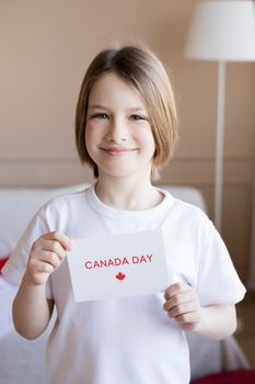 A young child smiling and holding a Canada Day sign indoors, representing Canadian patriotism.
