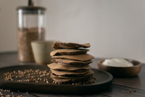 Healthy buckwheat pancakes on plate with scattered grains placed on wooden table with sour cream in light kitchen at home