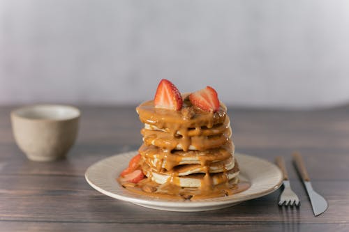Appetizing pancakes topped with ripe strawberries and sweet caramel served on wooden table with cutlery and mug in light kitchen