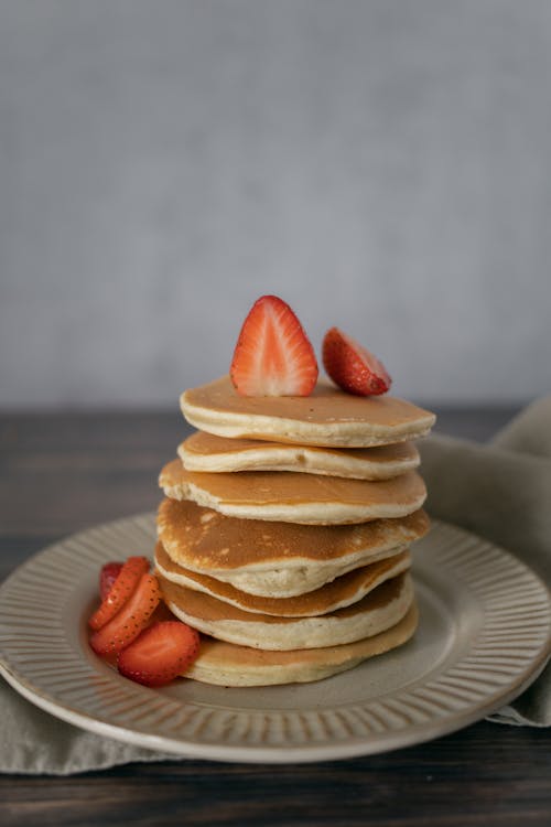 Heap of delicious pancakes served with cut fresh strawberries on white plate placed on wooden table with napkin in light kitchen
