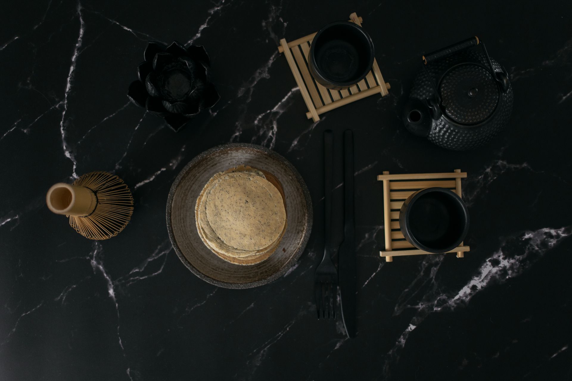 Top view stack of pancakes served in bowl amidst bamboo whisk and black mugs placed on marble surface in room