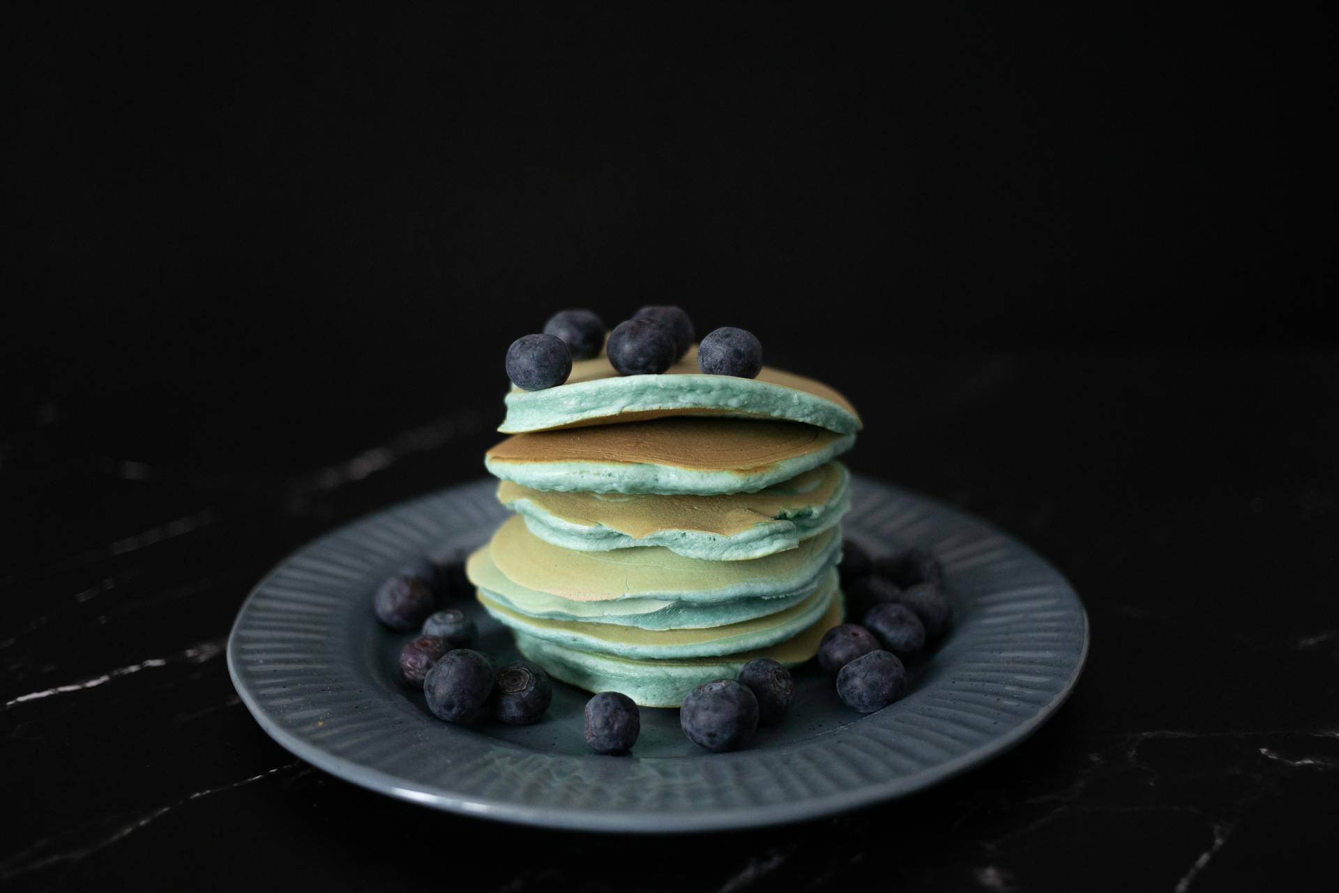 Heap of appetizing blue pancakes topped with ripe blueberries served on round plate with scattered berries against black background in studio