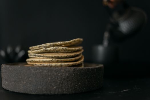 Heap of fresh pancakes served in bowl placed in table with kettle and cup on blurred black background during tea time
