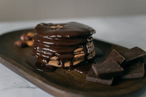 Delicious sweet pancakes and melted chocolate on top on wooden board with bar pieces on white table