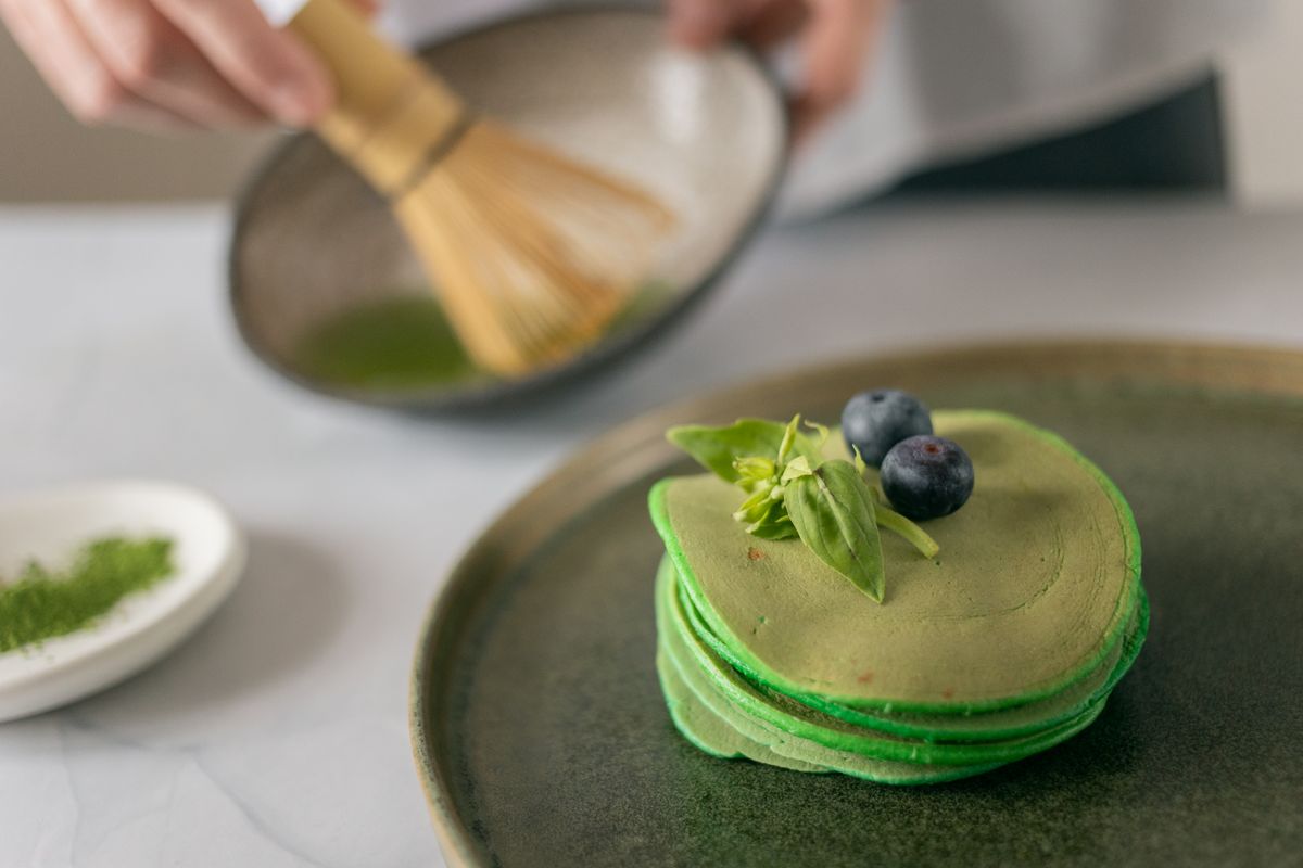Bamboo whisk stirring green batter beside a pancake stack topped with blueberries