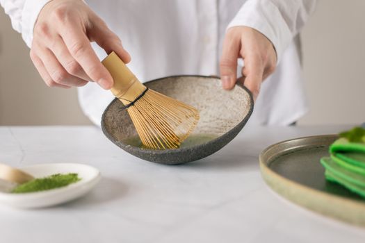 Crop unrecognizable cook in white shirt mixing dry food coloring with water in bowl using whisk on table with green pancakes