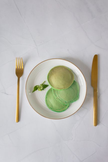 Top view of white plate with green colored pancakes with leaves near fork and knife on white table