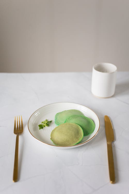 From above of tasty pancakes on white plate near cutlery and empty cup on white table in kitchen