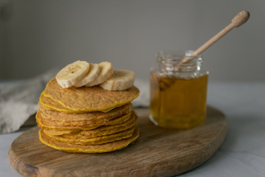 Tasty homemade pancakes with sliced bananas near jar with honey and wooden spoon on cutting board on table